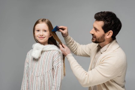 Foto de Happy bearded father brushing hair of smiling daughter isolated on grey - Imagen libre de derechos