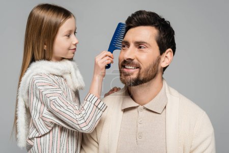 Foto de Happy girl brushing hair of smiling and bearded father isolated on grey - Imagen libre de derechos