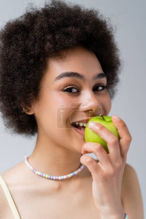 Foto de Sonriente mujer afroamericana mordiendo manzana aislada en gris - Imagen libre de derechos