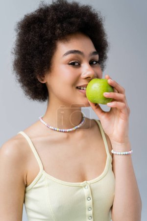 Retrato de la sonriente mujer afroamericana sosteniendo manzana verde aislada en gris 