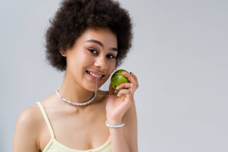 Fröhliche afrikanisch-amerikanische Frau hält frischen Apfel in der Hand und blickt isoliert in die Kamera auf grau 