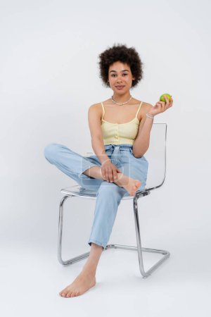 Barfuß afrikanische Amerikanerin hält frischen Apfel in der Hand, während sie auf einem Stuhl auf grauem Hintergrund sitzt 
