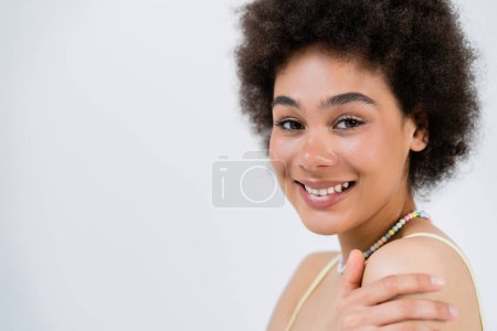 Positive african american woman touching shoulder and looking at camera isolated on grey 