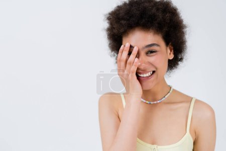Téléchargez les photos : Femme afro-américaine positive touchant le visage isolé sur le gris - en image libre de droit