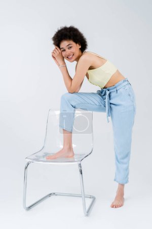 Full length of african american woman posing near modern chair on grey background 