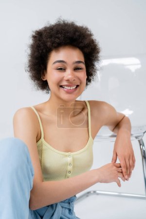 Photo for Cheerful african american model looking at camera near modern chair on grey background - Royalty Free Image