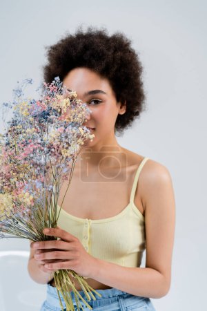 Mujer afroamericana sosteniendo flores cerca de la cara sobre fondo gris 