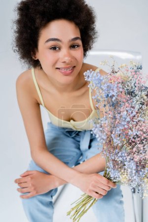 Smiling african american woman holding baby breath flowers while sitting on chair on grey background 