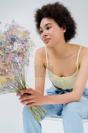 Junge afrikanisch-amerikanische Frau hält Blumen in der Hand, während sie auf einem Stuhl sitzt, isoliert auf grau 