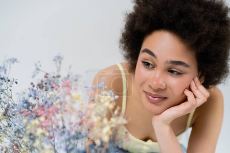Mujer americana bastante africana mirando flores borrosas aliento de bebé aisladas en gris 