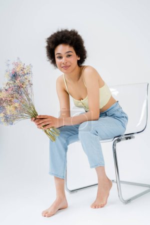 Barefoot woman in pants and top holding flowers while sitting on chair on grey background 