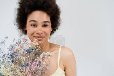 Lächelnde Afroamerikanerin blickt in die Kamera in der Nähe von Baby Atem Blumen isoliert auf grau 