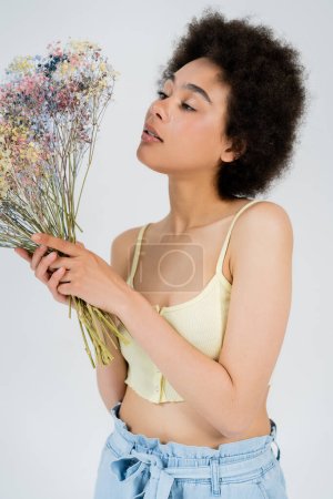 Jolie femme afro-américaine en haut tenant des fleurs de souffle de bébé isolées sur gris 