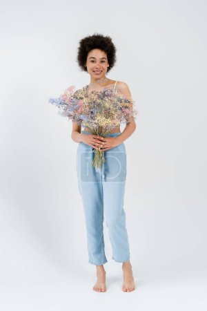 Full length of barefoot african american woman holding baby breath flowers on grey background 