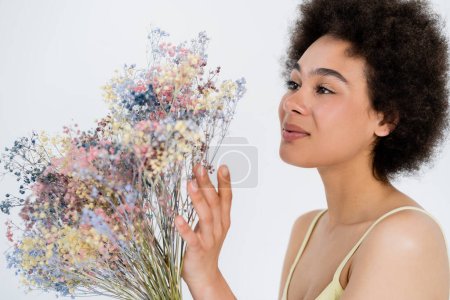 Agradable mujer afroamericana tocando flores aliento bebé aislado en gris 