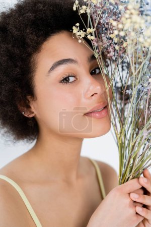 Retrato de una mujer afroamericana muy bonita sosteniendo flores de aliento de bebé aisladas en gris 