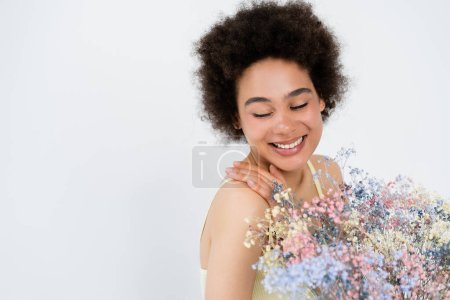 Happy african american woman touching shoulder near baby breath flowers isolated on grey, banner 