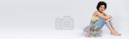 African american model in top and pants sitting near dry flowers on grey background, banner 