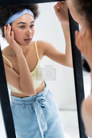 Hübsche afrikanisch-amerikanische Frau mit Stirnband, das Gesicht in der Nähe eines Spiegels auf grauem Hintergrund berührt 