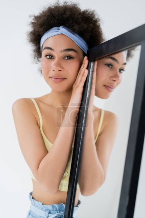 Smiling african american woman in headband looking at camera near mirror isolated on grey 