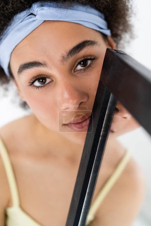Pretty african american woman with headband looking at camera near blurred mirror isolated on grey 