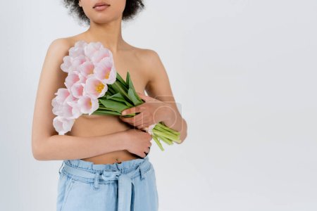Ausgeschnittene Ansicht einer jungen afrikanisch-amerikanischen Frau mit nackten Schultern, die Blumen isoliert auf grau hält 