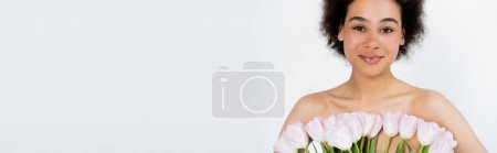 Petty african american woman with naked shoulders posing with tulips isolated on grey, banner 