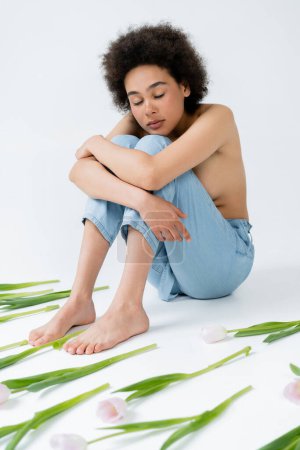 Young african american model with naked shoulders hugging knees near tulips on grey background 