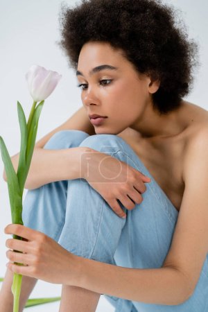 Modelo americano africano rizado con hombros desnudos sosteniendo tulipán mientras está sentado sobre fondo gris 