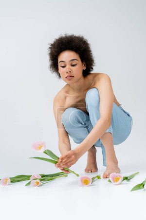 Descalza mujer afroamericana con hombros desnudos tomando tulipán sobre fondo gris 