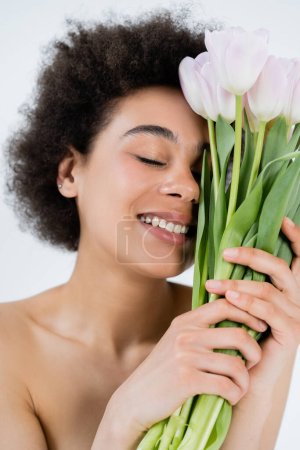 Photo for Smiling african american woman with naked shoulders holding bouquet of tulips isolated on grey - Royalty Free Image