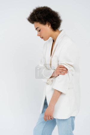 Foto de Vista lateral de la mujer afroamericana de moda enrollándose en la manga de la chaqueta aislada en gris - Imagen libre de derechos