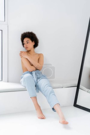 Hemdlos und barfuß sitzt eine Afroamerikanerin auf der Fensterbank vor grauem Hintergrund 
