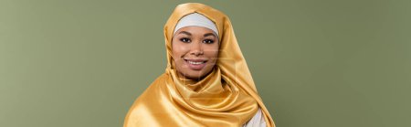 Portrait of smiling multiracial woman in yellow hijab looking at camera isolated on green, banner  