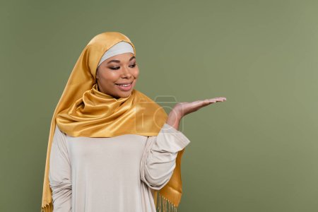 Positive multiracial woman in hijab pointing with hand isolated on green 