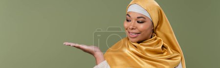 Positive multiracial woman in golden hijab pointing with hand isolated on green, banner 