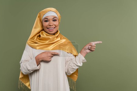 Carefree multiracial woman in hijab pointing with fingers isolated on green 