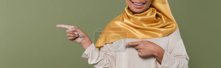 Vue recadrée de la femme souriante multiraciale en hijab pointant les doigts isolés sur vert, bannière 