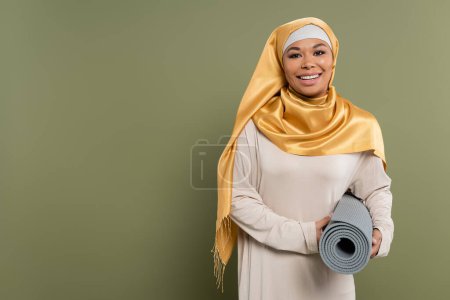 Sonriente mujer multirracial en hijab celebración de alfombra de fitness sobre fondo verde