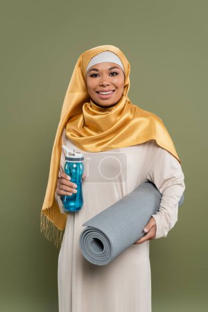 Carefree multiracial woman in hijab holding fitness mat and sports bottle isolated on green 