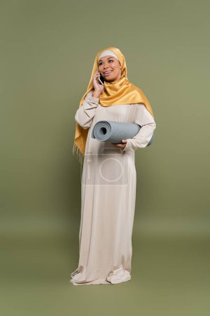 Full length of multiracial woman in hijab holding fitness mat and talking on smartphone on green background