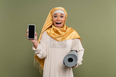 Mujer multirracial emocionada en hijab sosteniendo teléfono inteligente y alfombra de fitness sobre fondo verde