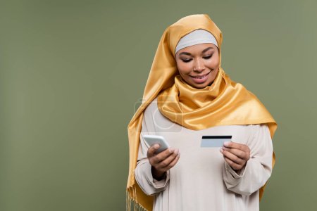 Photo pour Sourire femme multiraciale dans le hijab en utilisant smartphone et carte de crédit isolé sur vert - image libre de droit