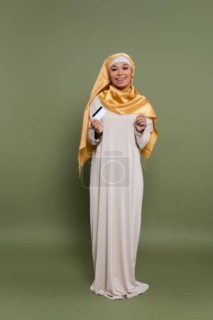 Full length of smiling multiracial woman in hijab holding credit card on green background