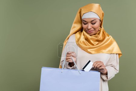 Multiracial woman in hijab putting credit card in shopping bag isolated on green 