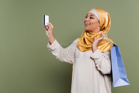 Mujer alegre multirracial en hijab dorado sosteniendo tarjeta de crédito y bolsa de compras sobre fondo verde