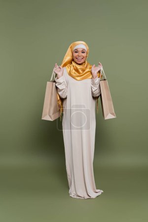 Positive multiracial woman in hijab holding shopping bags on green background