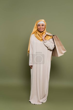 Photo pour Pleine longueur de souriante femme multiraciale dans le hijab tenant des sacs à provisions sur fond vert - image libre de droit
