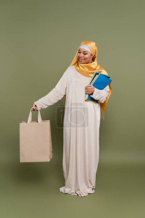 Carefree multiracial student in hijab holding shopping bags and notebooks on green background