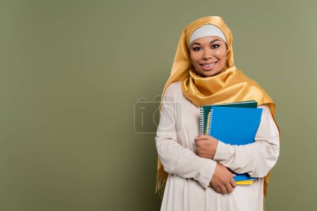 Cheerful multicultural student in hijab holding notebooks on green background